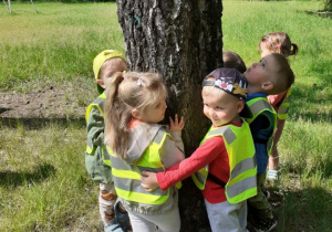 Na zdjęciu grupa dzieci przytula się do drzewa.