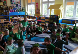 Zdjęcie grupowe w klasie na dywanie - dzieci wraz z nauczycielką. Wszyscy w zielonych strojach i z papierowymi opaskami emblematem Żabek na głowach.