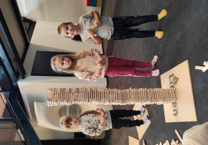 Dwie dziewczynki i chłopiec stoją obok wieży zbudowanej z drewnianych klocków.