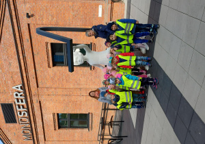 Zdjęcie grupowe dzieci i opiekunów stojących przy rzeźbie białej dziewczynki, na tle budynku z czerwonej cegły.