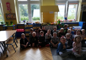 dzieci siedzą na podłodze za nimi wystawa z owoców przyniesionych przez dzieci