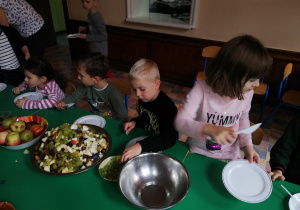 Dzieci na jadalni przy wspólnym stole kroją plastikowymi nożykami na małych talerzykach owoce na sałatkę