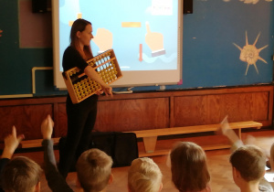 dzieci na sali przed tablica multimedialna, prowadzącą zajęcia objaśnia z pomocą projekcji na tablicy multimedialnej działanie japońskiego liczydła oraz zadaje dzieciom proste zagadki matematyczne
