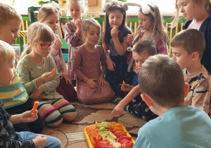 Zdjęcie przedstawia grupę dzieci siedzących na dywanie i jedzących owoce i warzywa.