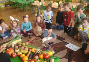 Na zdjęciu dzieci siedzą na dywanie i dzielą przyniesione owoce i warzywa na odpowiednie kategorie.