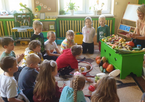 Na zdjęciu dzieci siedzą na dywanie i dzielą przyniesione owoce i warzywa na odpowiednie kategorie.
