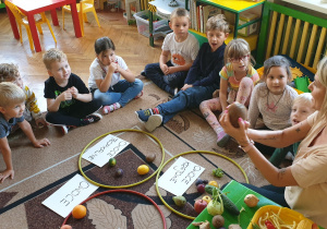 Na zdjęciu dzieci siedzą na dywanie a nauczycielka pokazuje im kokosa.