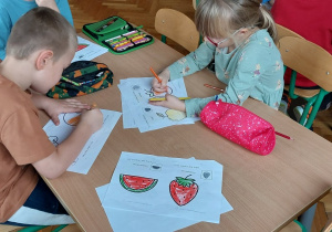 Zdjęcie przedstawia dzieci siedzące przy stoliku i kolorujące rysunki owoców.