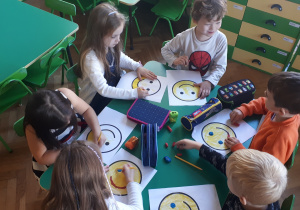 na zdjęciu dzieci siedzące przy stoliku wykonujące prace plastyczną z wykorzystaniem kredek i plasteliny