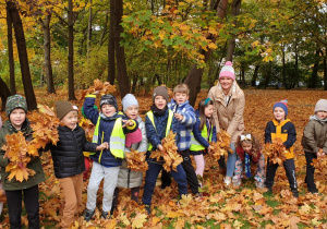 Na zdjęciu cała grupa z nauczycielką na tle jesiennych drzew przygotowuje się do rzucenia liści.