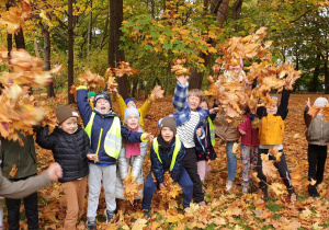 Na zdjęciu cała grupa z nauczycielką na tle jesiennych drzew rzuca do góry liście.