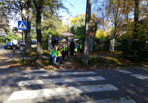 Na zdjęciu dzieci stoją ubrane w kamizelki odblaskowe przed przejściem dla pieszych