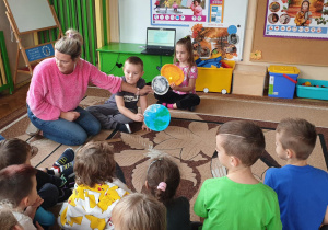 Na zdjęciu dzieci siedzą na dywanie i obserwują jak nauczycielka pokazuje i tłumaczy schemat powstawania zaćmienia.