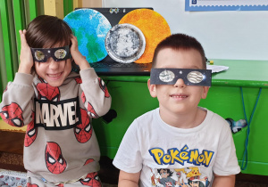 Na zdjęciu dwaj chłopcy przymierzający specjalne okulary do oglądania zjawiska zaćmienia słońca.