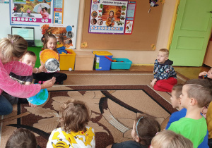 Na zdjęciu dzieci siedzą na dywanie i obserwują jak nauczycielka pokazuje i tłumaczy schemat powstawania zaćmienia.