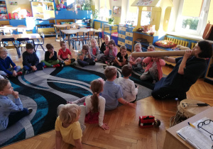 Grupa dzieci siedzi w klasie na dywanie i przygląda się mamie Tadzia, która pokazuje, jak wydobyć dźwięk z długiej , papierowej tuby