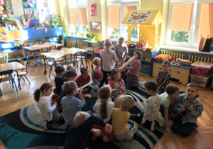Na zdjęciu grupa dzieci siedząc na dywanie w klasie ogląda i gra na instrumentach muzycznych