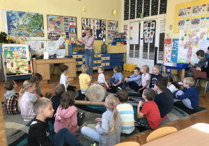Na zdjęciu grupa dzieci siedząc na dywanie w klasie słucha i przygląda się nauczycielce grającej na flecie poprzecznym