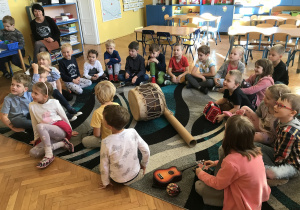 Na zdjęciu grupa dzieci siedząc na dywanie w klasie ogląda i gra na instrumentach muzycznych