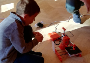 Na zdjęciu chłopiec kuca przy małej drukarce 3D, trzyma w ręce materiał do drukowania w 3D.