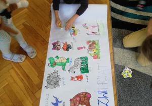 dzieci naklejają na duży arkusz białego papieru wycięte z papieru, pokolorowane obrazki różnych zwierząt