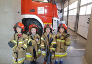 Dzieci w strojach strażackich pozują do zdjęcia. W tle widoczny samochodów strażacki.