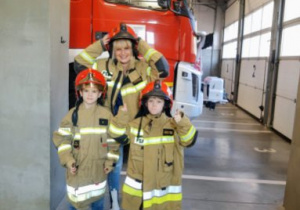 Dzieci w strojach strażackich pozują do zdjęcia. W tle widoczny samochodów strażacki.