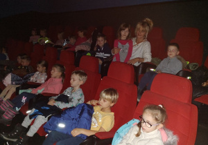 Na zdjęciu grupa dzieci siedzi na kinowych fotelach wraz z nauczycielką.