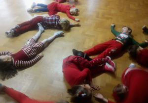 Dzieci ubrane na czerwono leżą na podłodze wykonując jedno z zadań podczas zabawy mikołajkowej.