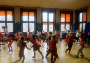 Dzieci w mikołajkowych strojach tańczą na sali gimnastycznej podczas mikołajkowej zabawy.