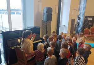 Dzieci stoją obok pianina, na którym gra Pani prowadząca warsztaty.