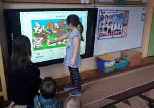 zdjęcie przedstawia dziewczynkę wraz z nauczycielką i częścią grupy podczas rozwiązywania zagadki wyświetlanej na monitorze