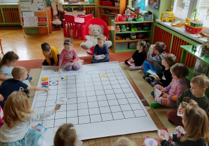 Dzieci siedzące wokół maty do kodowania. Układanie obrazków według kształtów i kolorów.