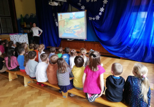 dzieci siedzą na ławeczkach , oglądają na tablicy multimedialnej prezentację o dinozaurach, o której opowiada nauczycielka stojąca przy tablicy