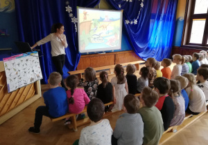 dzieci siedzą na ławeczkach , oglądają na tablicy multimedialnej prezentację o dinozaurach, o której opowiada nauczycielka stojąca przy tablicy