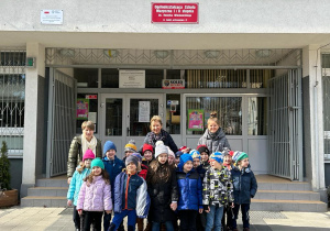 Dzieci wraz z nauczycielkami stoją przed budynkiem OSM I i II st. w Łodzi, w tle widać wejście do szkoły oraz tabliczkę z nazwa szkoły