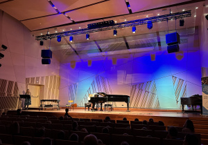 Na zdjęciu podświetlona kolorowo scena sali koncertowa z fortepianem na którym gra duet młodych muzyków.