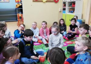 dzieci siedzą na kolorowej macie i grają na małych instrumentach perkusyjnych