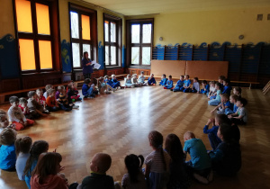 dzieci siedzą w ogromnym kole na podłodze na sali, w tle nauczycielka czyta zagadki