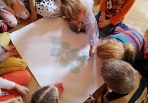 Dzieci siedzą na podłodze i układają papierowe kropelki na kartonie tworząc obrazek