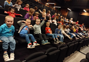 Na zdjęciu dzieci siedzą uśmiechnięte na widowni w teatrze, czekając na przedstawienie.