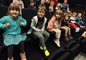 Na zdjęciu dzieci siedzą uśmiechnięte na widowni w teatrze, czekając na przedstawienie.