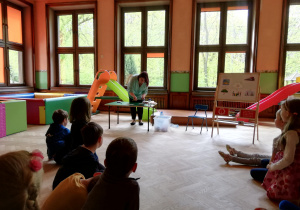 dzieci siedzą w półkolu, prowadzącą pokazuje przy stoliku doświadczenie z kolorową kroplą wody rozdmuchaną słomką na kartce papieru