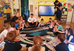 Grupa dzieci siedzi na dywanie, przed sobą dzieci trzymają rysunki z plecaczkami, wśród dzieci siedzi wiceprezydent Łodzi, który omawia z dziećmi ich rysunki.