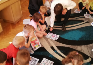 Grupa dzieci siedzi na dywanie, przed sobą dzieci trzymają rysunki z plecaczkami, wśród dzieci siedzi wiceprezydent Łodzi, który omawia z dziećmi ich rysunki.