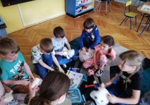 Dzieci siedzą na dywanie i pod kierunkiem studenta medycyny uczą się używając zabawkowych narzędzi medycznych badać swoje maskotki misiów, które są ich pacjentami.