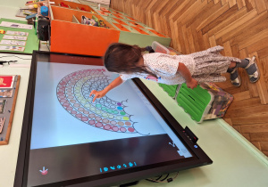 Dzieci kolorują kropkową tęczę na tablicy multimedialnej.