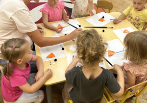 Na zdjęciu grupa dzieci wraz z nauczycielką wykonują przy stoliku pracę plastyczną.