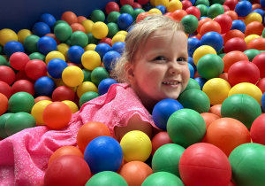Na zdjęciu uśmiechnięta dziewczynka bawi się w basenie z kolorowymi, plastikowymi piłeczkami.