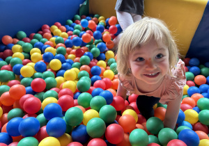 Na zdjęciu uśmiechnięta dziewczynka bawi się w basenie z kolorowymi, plastikowymi piłeczkami.